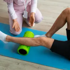 Fizjoterapeuta pomaga pacjentowi w ćwiczeniach po endoprotezie kolana, używając wałka do masażu.
