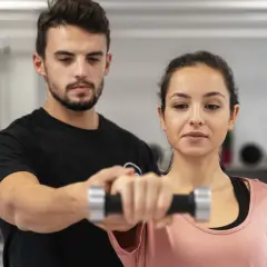 Trener pomaga kobiecie ćwiczyć z hantlem. To przykład, jak fitness co to jest – aktywność fizyczna dla zdrowia i lepszego samopoczucia.