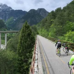 Rowerzyści przemierzają malowniczą trasę **Alpe Adria** w otoczeniu gór i zielonych lasów. Widoczny most i rzeka dodają krajobrazowi majestatu.