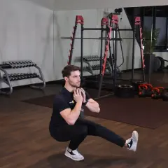 Mężczyzna wykonuje ćwiczenie pistol squat z hantlem. W tle siłownia z maszynami i ciężarami.