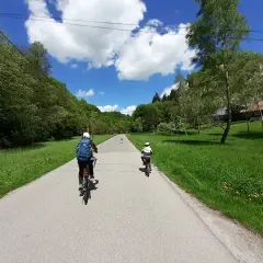 Dwie osoby na rowerach jadą malowniczą trasą rowerową w Ojcowie. W tle zielone wzgórza i błękitne niebo z chmurami.