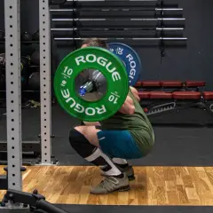 Mężczyzna wykonuje głęboki **squat** z ciężarem na plecach w siłowni. Zielone talerze Rogue ważą 10 kg każdy.