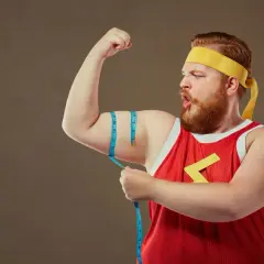 Mężczyzna typu endomorfik z wąsami i brodą, w żółtej opasce i czerwonej koszulce, mierzy obwód bicepsa.