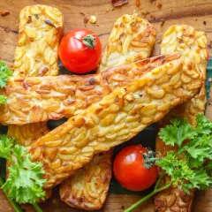 Usmażony tempeh, czyli co to jest? Złociste kawałki na drewnianej desce, ozdobione pomidorkami i natką pietruszki.