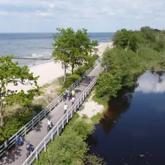 Ludzie na rowerach przemierzają malowniczą ścieżkę Velo Baltica, z jednej strony morze i plaża, z drugiej spokojna, ciemna woda.