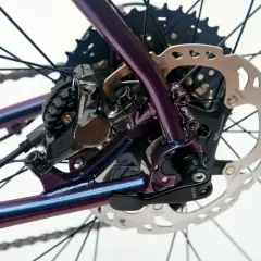 Błyszcząca, fioletowa rama roweru z widocznym hamulcem tarczowym Shimano i kasetą. Sztywna oś w rowerze zapewnia stabilność.