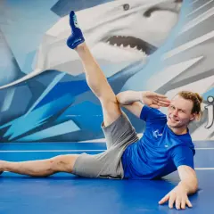 Mężczyzna w niebieskiej koszulce i szarych spodenkach wykonuje ćwiczenie rozciągające na niebieskiej macie.