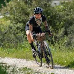 Kobieta z tatuażami na rowerze, ciesząca się kolarstwem gravelowym na piaszczystej drodze wśród zieleni.