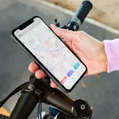 Dłoń trzyma smartfon z mapą i ikonami aplikacji rowerowych, oparty o kierownicę roweru.