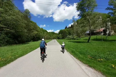 Dwie osoby na rowerach jadą malowniczą trasą rowerową w Ojcowie. W tle zielone wzgórza i błękitne niebo z chmurami.