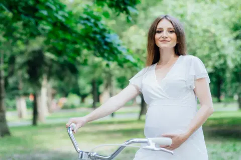 Uśmiechnięta ciężarna kobieta w białej sukience trzyma kierownicę roweru w parku.