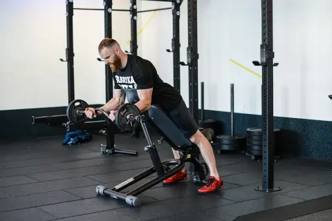 Mężczyzna z brodą wykonuje spider curl na ławce treningowej, podnosząc sztangę.
