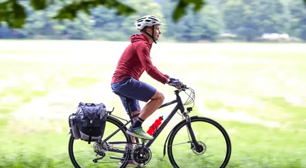 Mężczyzna w kasku jedzie na rowerze trekkingowym z sakwami, ciesząc się przejażdżką pośród zieleni.