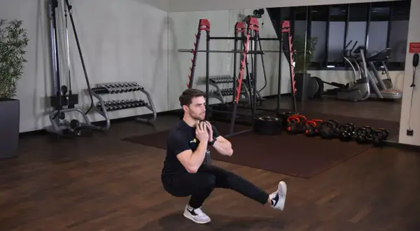 Mężczyzna wykonuje ćwiczenie pistol squat z hantlem. W tle siłownia z maszynami i ciężarami.