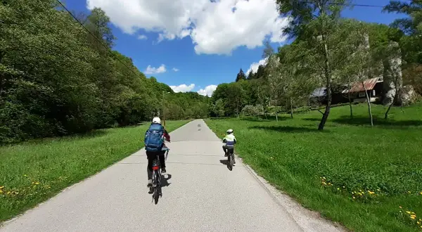 Dwie osoby na rowerach jadą malowniczą trasą rowerową w Ojcowie. W tle zielone wzgórza i błękitne niebo z chmurami.