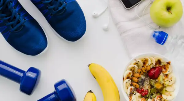 Niebieskie buty sportowe, hantle, miska owsianki z owocami, jabłko, telefon i butelka wody.