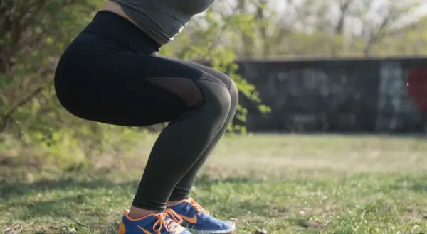 Kobieta w czarnych legginsach i sportowym topie wykonuje przysiady na trawie.