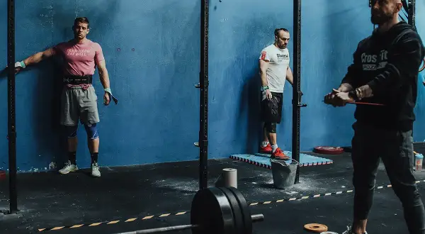 Trzech mężczyzn w siłowni CrossFit, jeden oparty o ścianę, drugi przy stojaku, trzeci z gumą treningową.