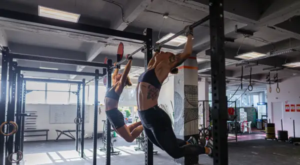 Dwie kobiety wykonują podciąganie na drążku w siłowni. To część treningu, który może być elementem "fran crossfit co to jest".