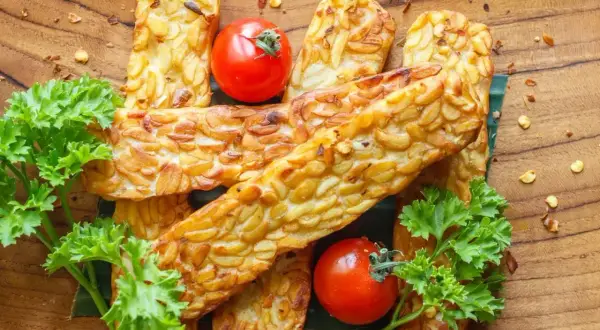 Usmażony tempeh, czyli co to jest? Złociste kawałki na drewnianej desce, ozdobione pomidorkami i natką pietruszki.