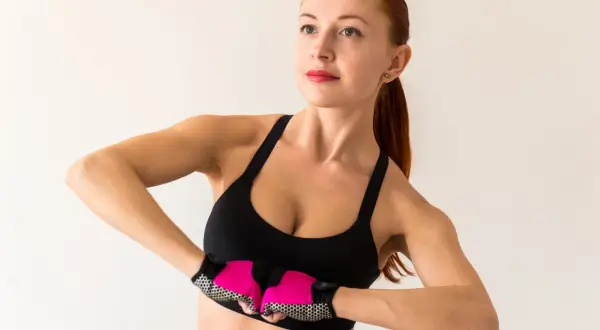 Kobieta w sportowym topie wykonuje ćwiczenia na podniesienie biustu, z różowymi rękawiczkami na dłoniach.