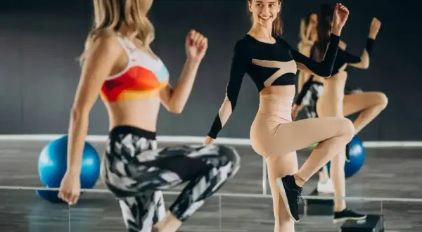Dwie kobiety w sportowych strojach wykonują tbc ćwiczenia na stepach. W tle niebieska piłka gimnastyczna.