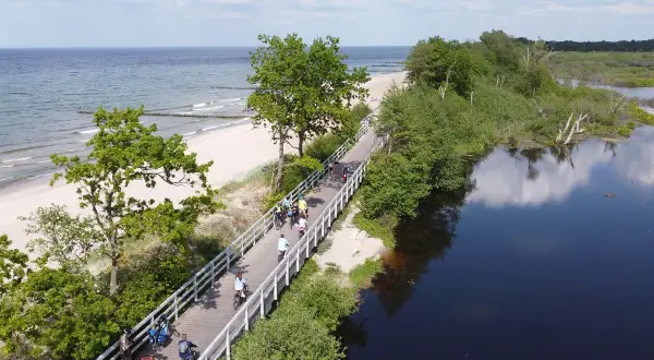Ludzie na rowerach przemierzają malowniczą ścieżkę Velo Baltica, z jednej strony morze i plaża, z drugiej spokojna, ciemna woda.