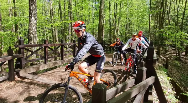 Grupa rowerzystów pokonuje drewniany mostek na leśnej trasie. Słońce przebija się przez liście, tworząc malowniczy krajobraz.