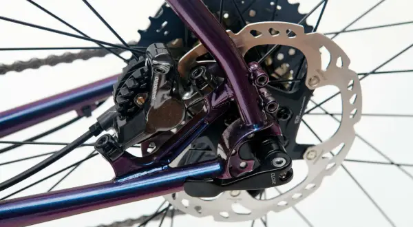 Błyszcząca, fioletowa rama roweru z widocznym hamulcem tarczowym Shimano i kasetą. Sztywna oś w rowerze zapewnia stabilność.