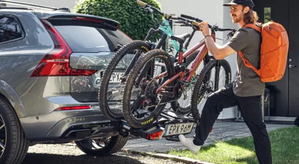 Mężczyzna z pomarańczowym plecakiem mocuje rowery do bagażnika. Widoczna tablica rejestracyjna na bagażniku rowerowym.