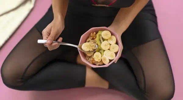 Kobieta w stroju sportowym je owsiankę z bananem i kiwi. Zastanawia się, czy lepiej zjesc przed czy po treningu.