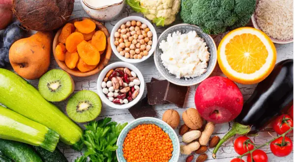 Kolorowy stół pełen zdrowych produktów: cukinia, kiwi, pomidory, bakłażan, jabłko, pomarańcza, brokuły, kalafior, soczewica, ciecierzyca, fasola, orzechy, migdały, suszone morele, ryż, twaróg i gorzka czekolada – idealne warzywa o niskim indeksie glike...