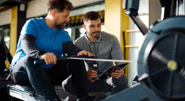 Trener crossfit omawia plan treningowy z podopiecznym na wioślarzu. Zastanawiasz się, ile zarabia trener crossfit?