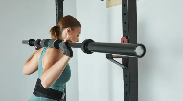 Kobieta w rękawiczkach i pasie treningowym przygotowuje się do przysiadów ze sztangą. Trening push pull legs to jej sposób na siłę.