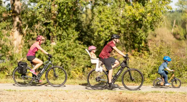 Rodzina na rowerach cieszy się aktywnym wypoczynkiem. Mama z bagażem, tata z córką w foteliku i mały chłopiec na swoim rowerze – idealny przykład turystyki rowerowej.