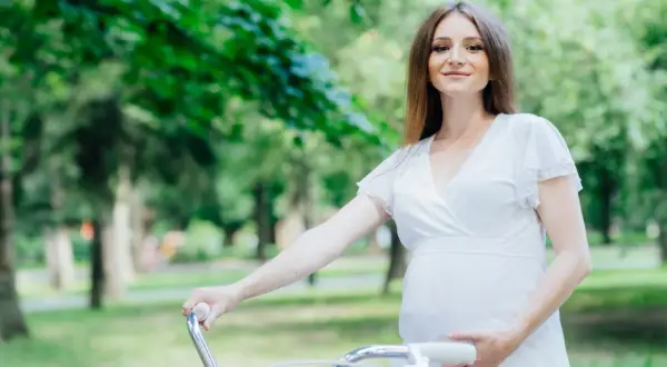 Uśmiechnięta ciężarna kobieta w białej sukience trzyma kierownicę roweru w parku.