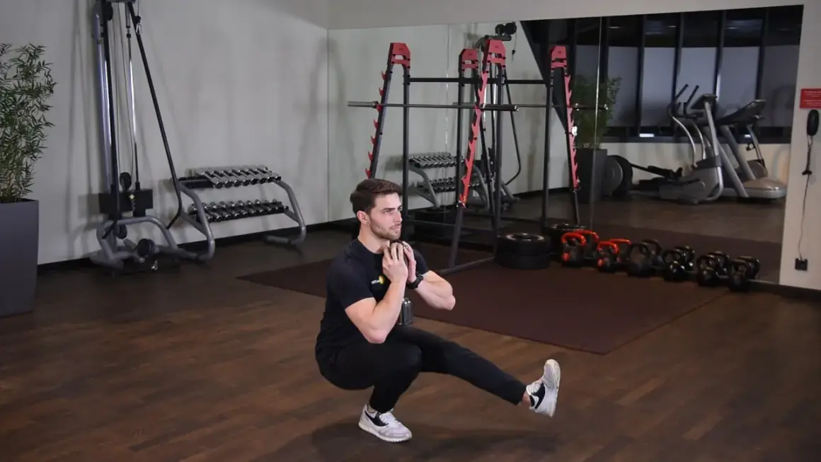 Mężczyzna wykonuje ćwiczenie pistol squat z hantlem. W tle siłownia z maszynami i ciężarami.