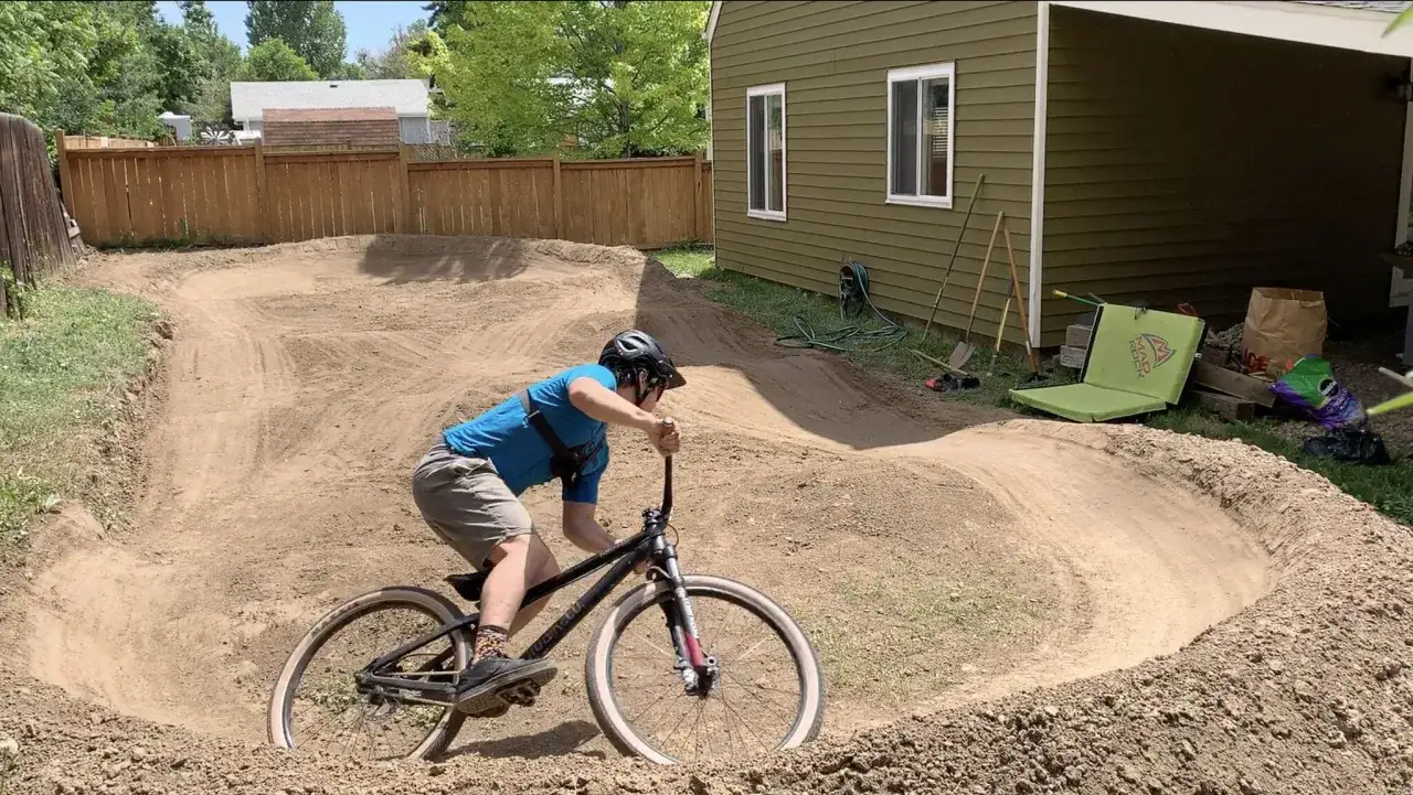 Chłopiec na rowerze testuje nowy pump track w ogrodzie. W tle dom i narzędzia ogrodnicze.