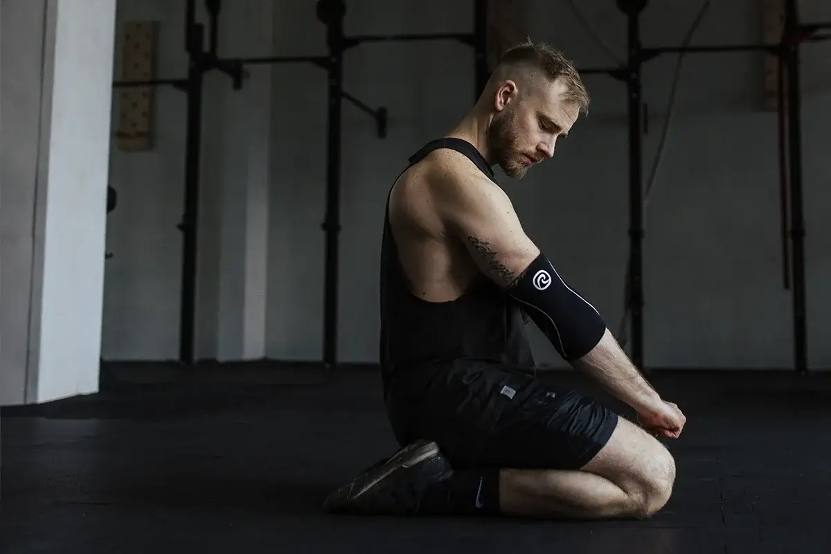 Mężczyzna w opasce na łokieć klęczy na siłowni. Czy crossfit jest zdrowy? To pytanie zadaje sobie wielu sportowc&oacute;w.