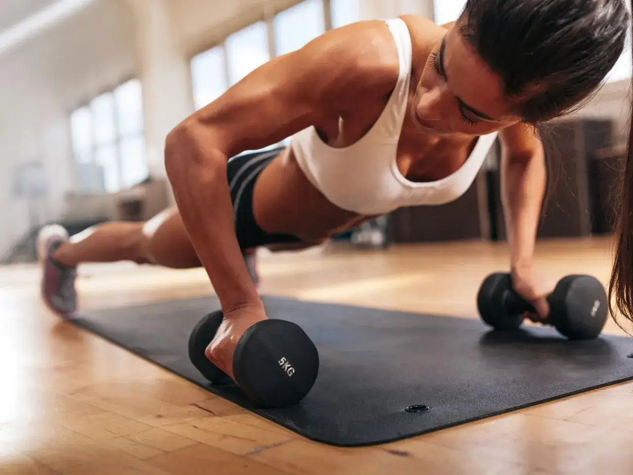 Kobieta wykonuje pompki z hantlami, ćwicząc siłę. To świetny spos&oacute;b, jak trenować crossfit w domu.