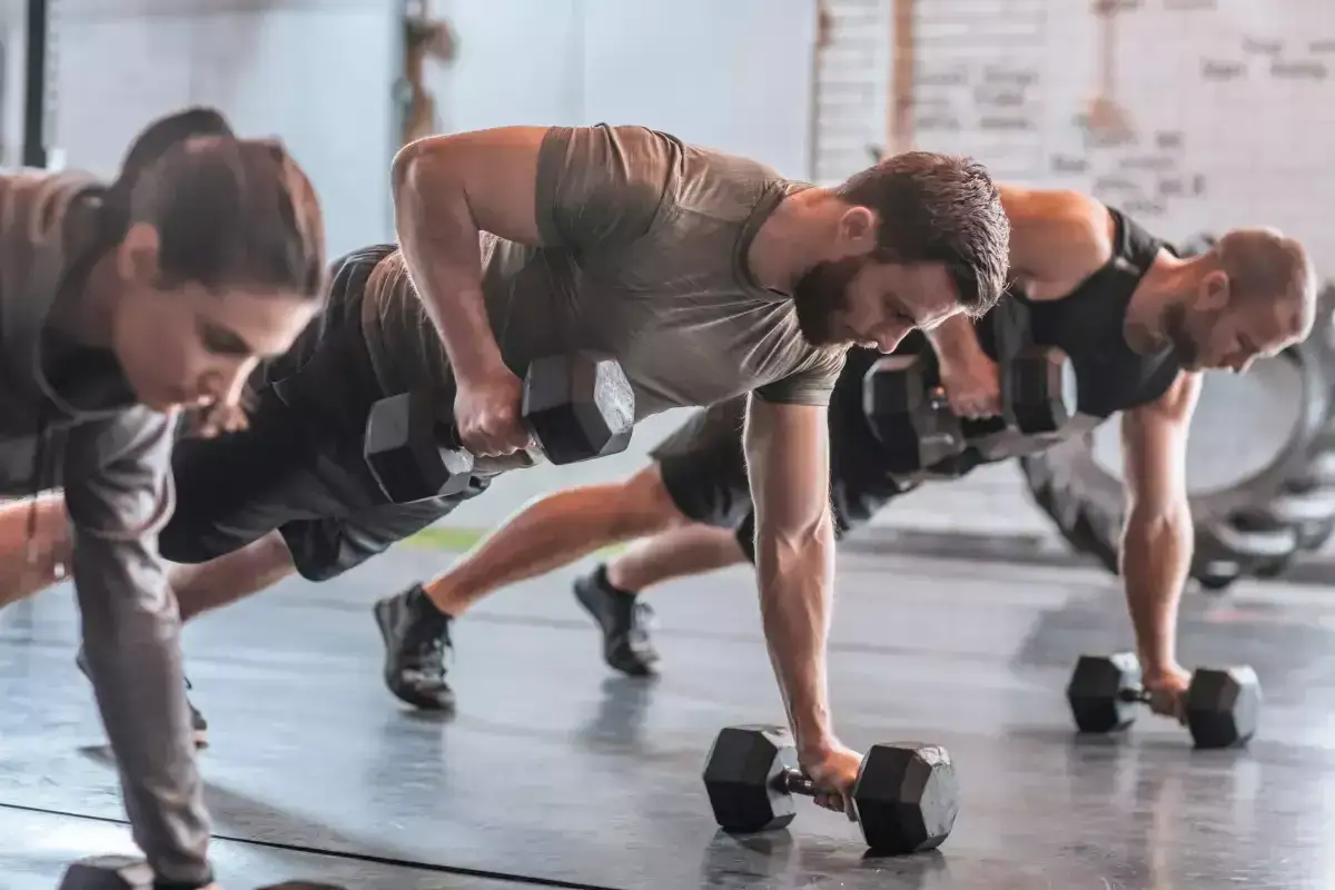 Ludzie wykonują ćwiczenia z hantlami w pozycji deski. To pokazuje, czym jest crossfit – trening siłowy i wytrzymałościowy.