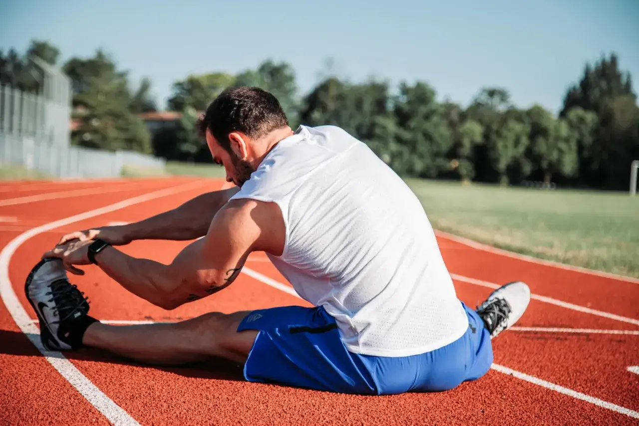 Mężczyzna w białej koszulce i niebieskich spodenkach wykonuje stretching na bieżni.