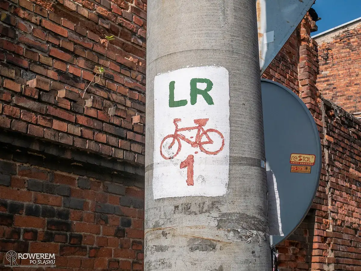 Znak rowerowy na słupie informuje o trasie LR 1. To początek przygody po czerwonym szlaku, gdzie ceglane mury opowiadają historie.