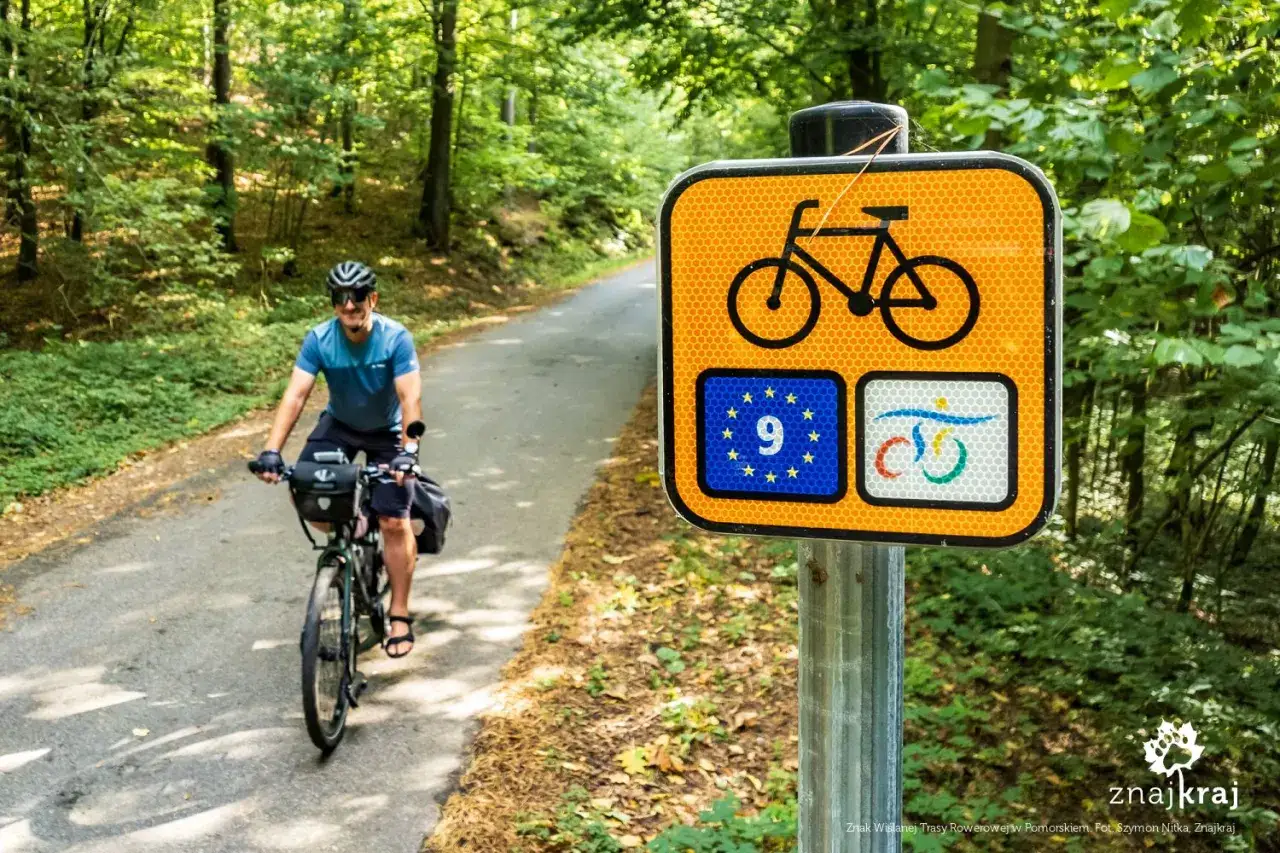 Mężczyzna na rowerze przemierza leśną ścieżkę. Obok drogi znak informuje o trasie rowerowej, zachęcając do turystyki rowerowej.