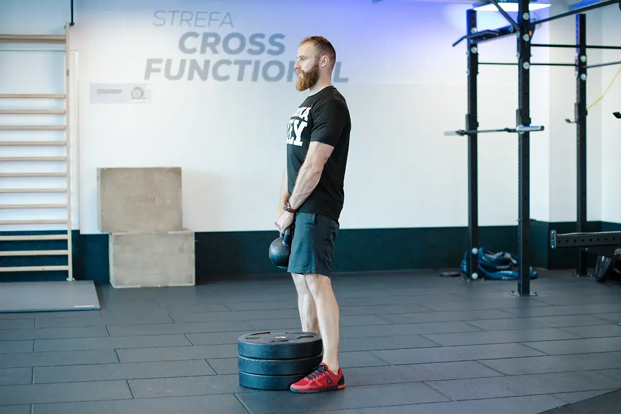 Mężczyzna wykonuje ćwiczenie rack pull z kettlebell, stojąc na stosie talerzy.