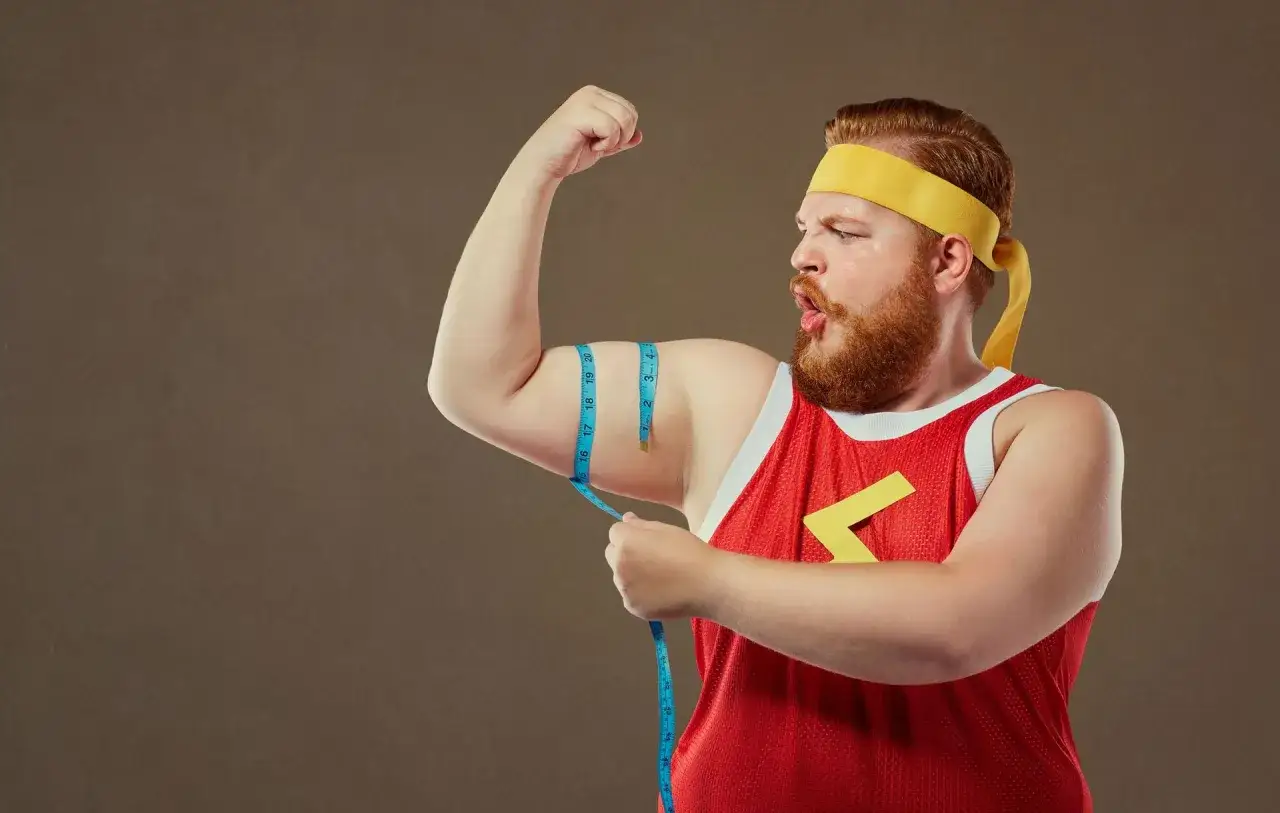 Rudowłosy mężczyzna typu endomorfik w żółtej opasce i czerwonej koszulce mierzy obwód bicepsa.