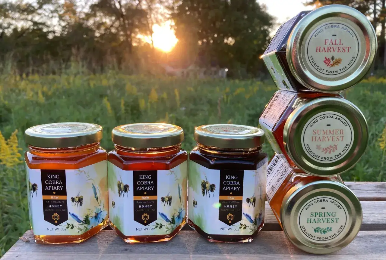 Słońce zachodzi nad łąką, oświetlając słoiki z miodem King Cobra Apiary. Czy miód jest zdrowy? Tak, ten naturalny, surowy miód prosto z ula to dowód!