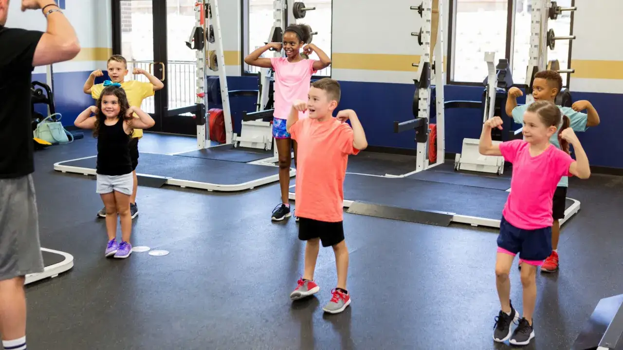 Dzieci ćwiczą w siłowni, pokazując bicepsy. Trening crossfit dla najmłodszych może być krótki i pełen zabawy.