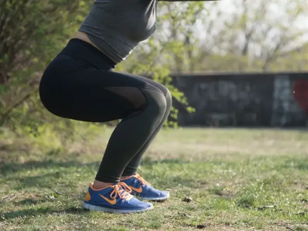 Kobieta w czarnych legginsach i sportowym topie wykonuje przysiady na trawie.