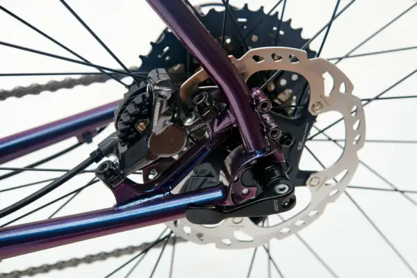 Błyszcząca, fioletowa rama roweru z widocznym hamulcem tarczowym Shimano i kasetą. Sztywna oś w rowerze zapewnia stabilność.